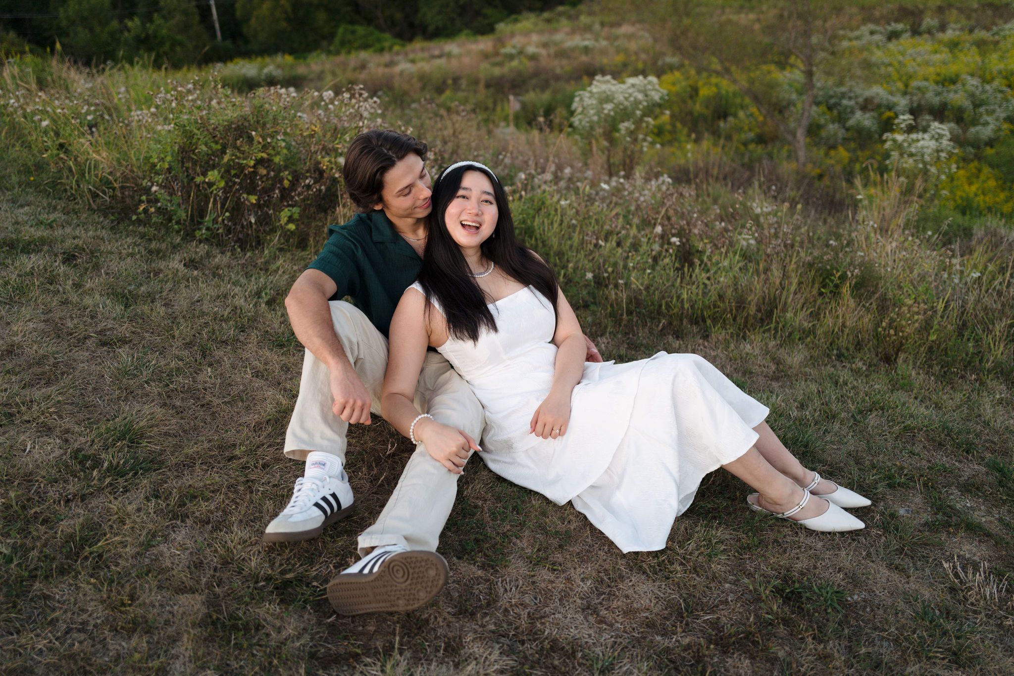 A couple in wedding attire sits together on a grassy hillside during sunset.