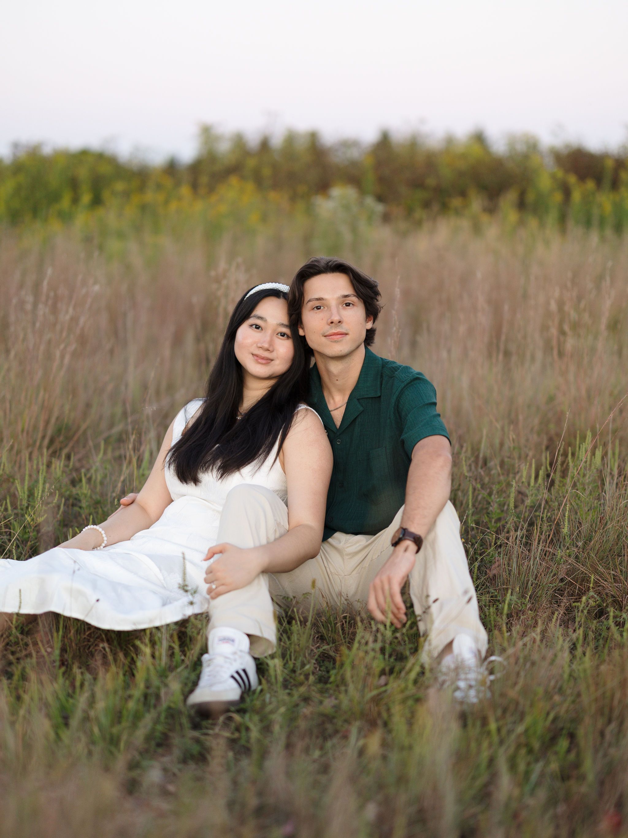 A couple in white and green attire sits together in a grassy meadow during golden hour.