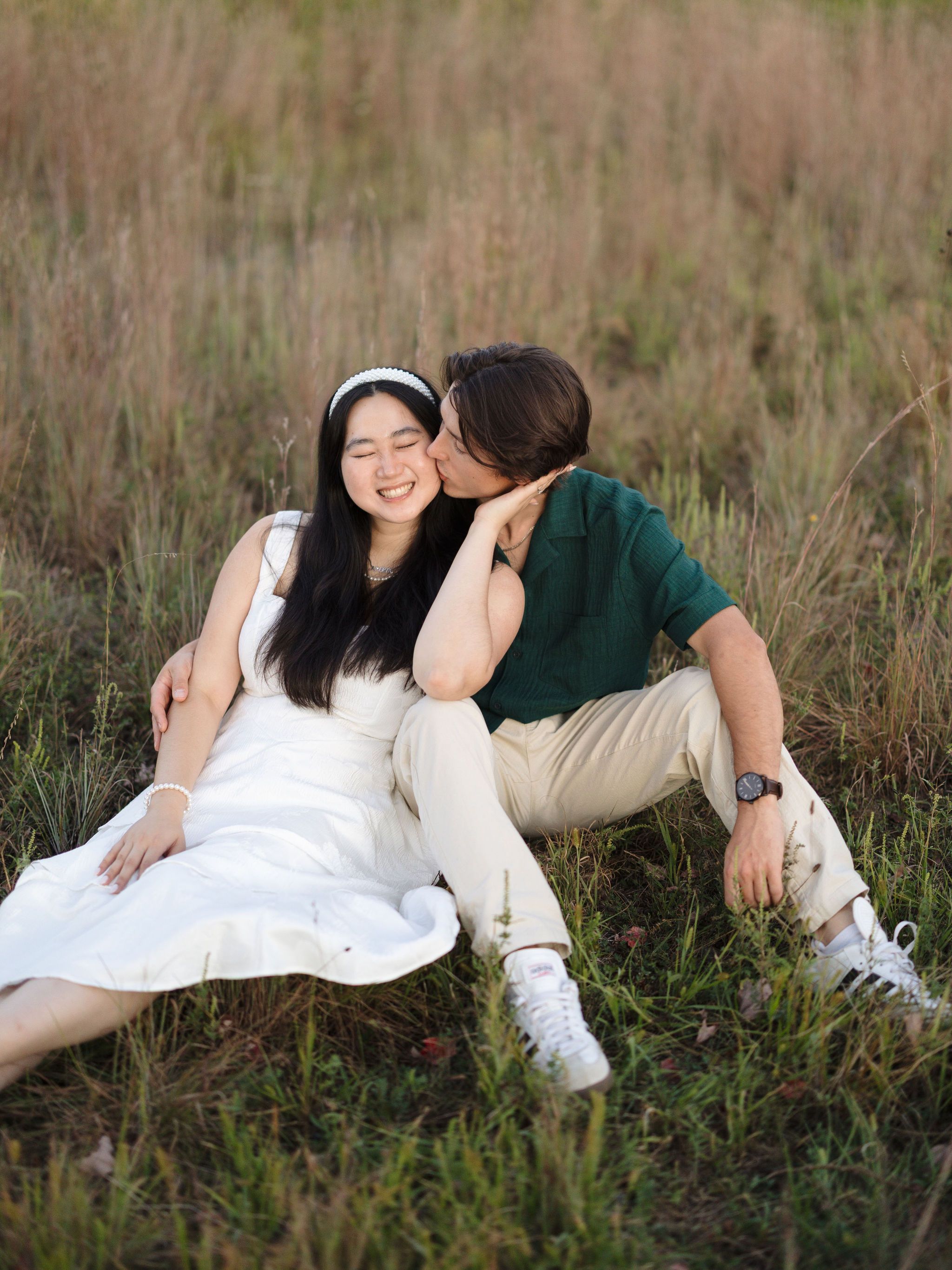 A couple in formal attire cuddles together while lying in a grassy field on a sunny day.