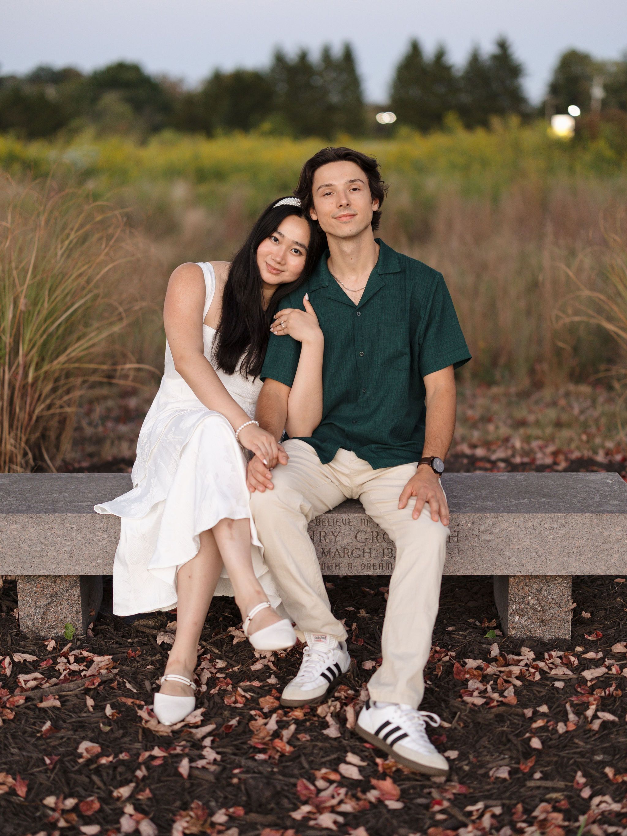 A couple sits outdoors wearing coordinated white and green outfits as autumn leaves scatter around wooden benches.
