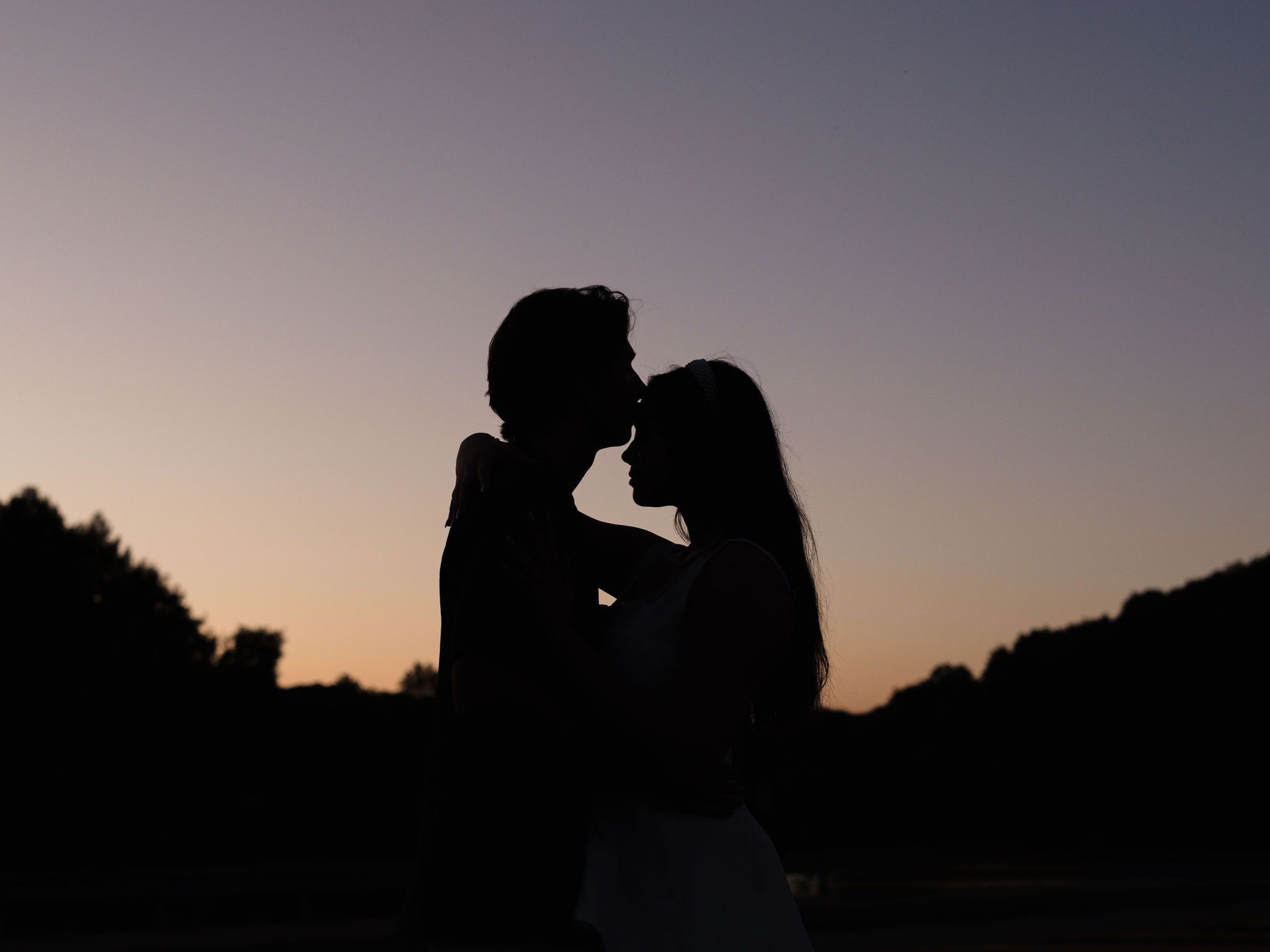 Two silhouettes stand close together against a sunset sky in a romantic scene.