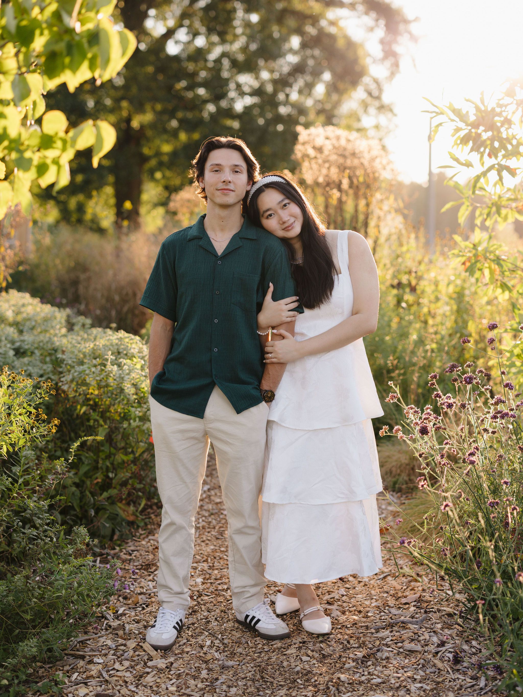 A couple poses together on a garden path at sunset with green foliage and natural lighting creating a romantic atmosphere.