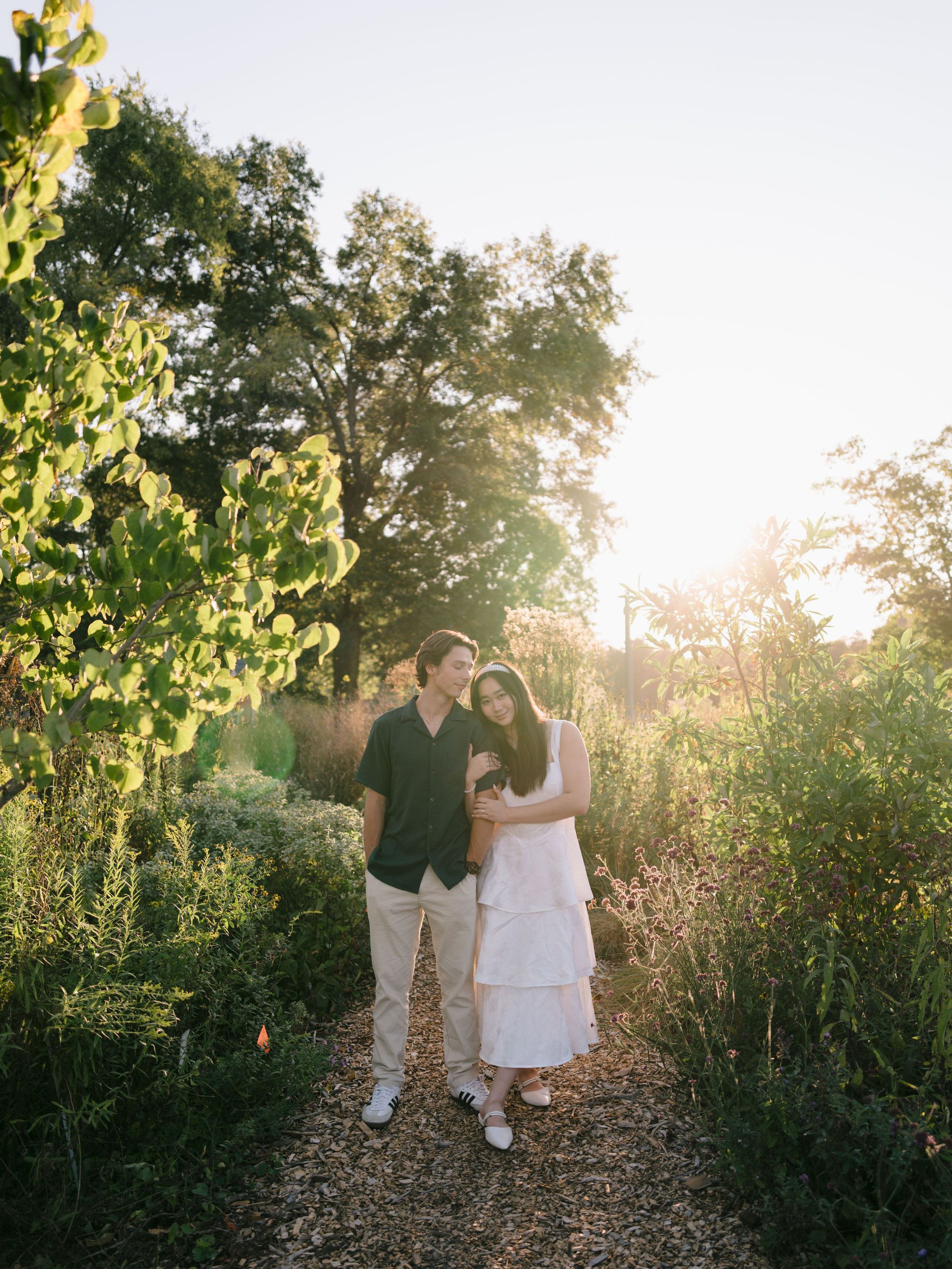 A romantic couple stands on a sunlit garden path surrounded by lush summer greenery and golden evening light.