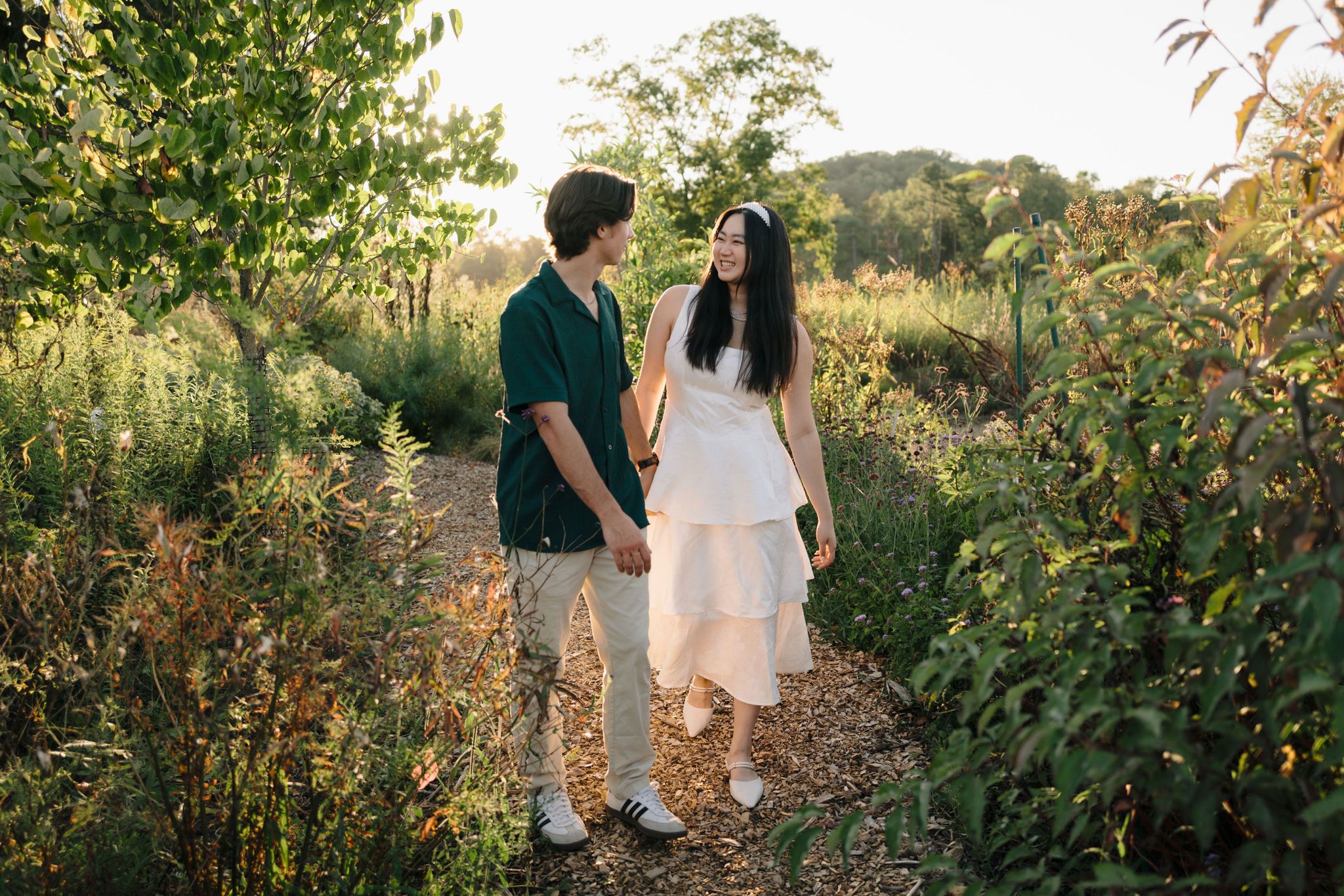 Couple holding hands walking along rustic garden path at sunset with golden light filtering through foliage.