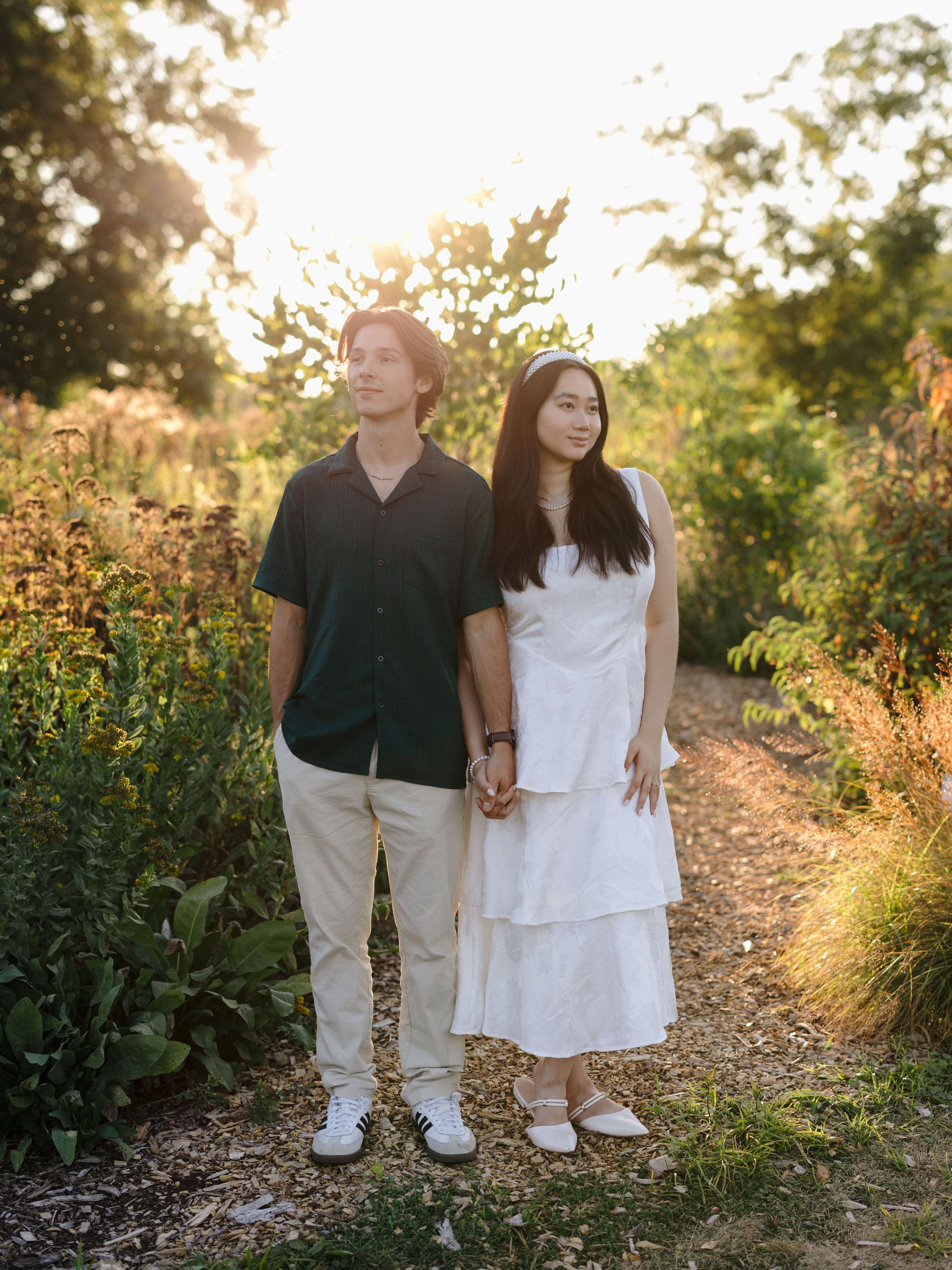 A couple holds hands while standing on a garden path at golden hour with sunlit foliage in the background.