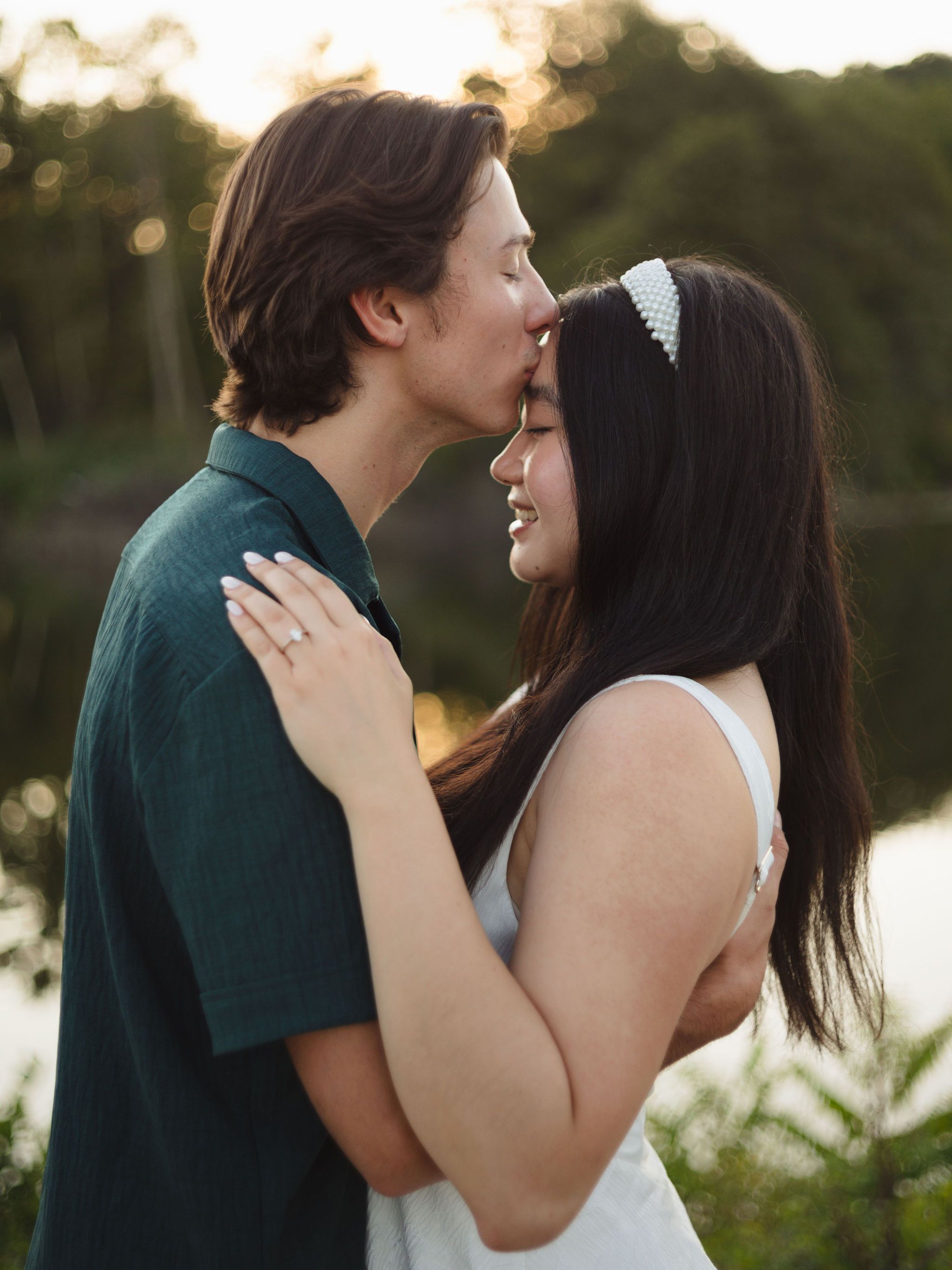 A tender romantic moment between a couple embracing in soft sunset lighting beside a lake.