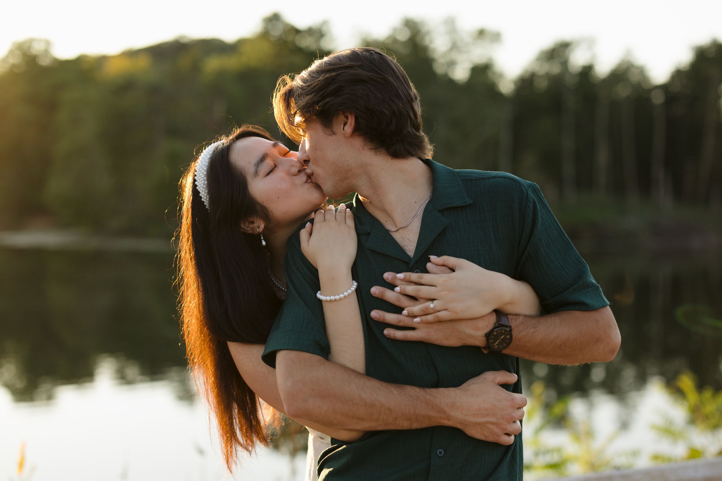 Couple in casual attire embraces romantically by a serene lake during sunset, creating a silhouette effect.
