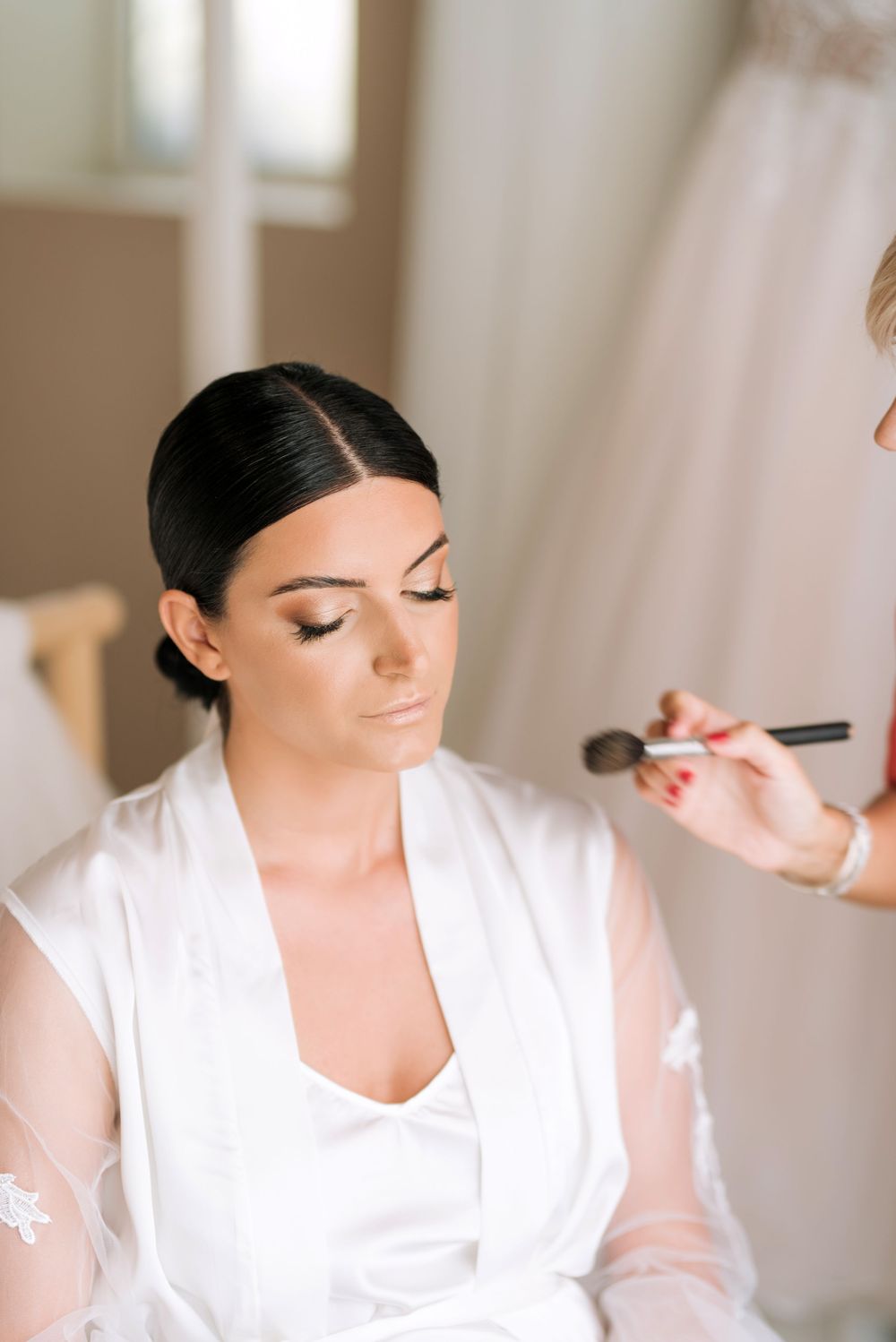 Professional makeup artist applies finishing touches and cosmetics during wedding day preparation in bright room.