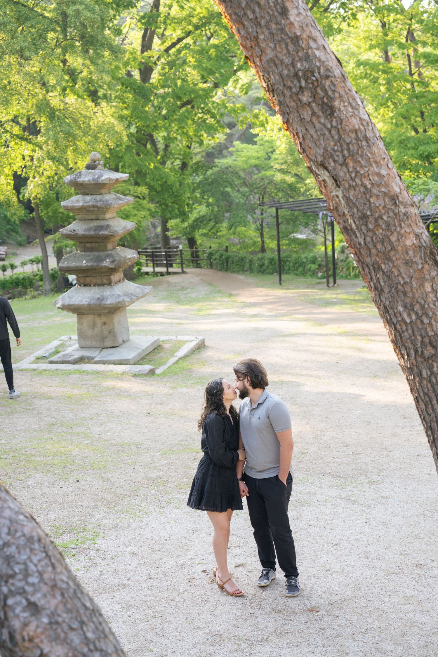 A couple stands near a stone lantern on a peaceful gravel path surrounded by trees in a tranquil garden setting.