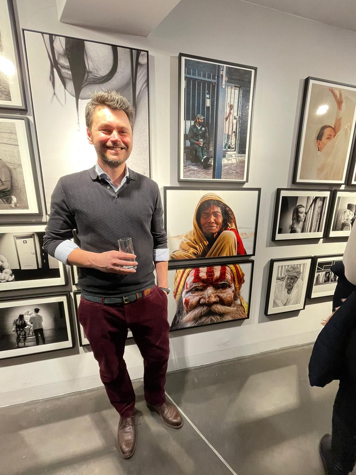 Person in burgundy pants sits casually on gallery floor surrounded by black and white photographs on white walls.