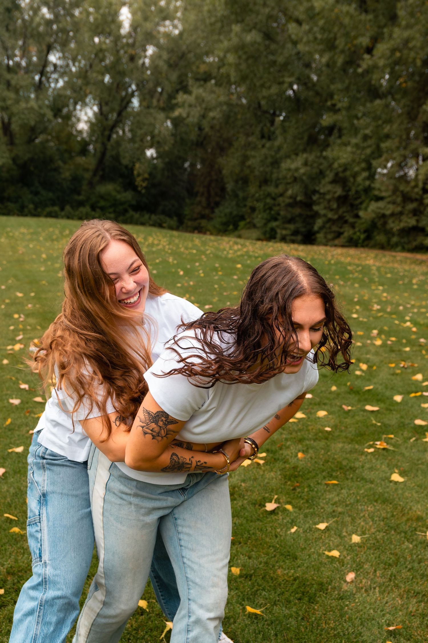 Two young women laughing and playing in a park, wearing white t-shirts and jeans on a fall day.