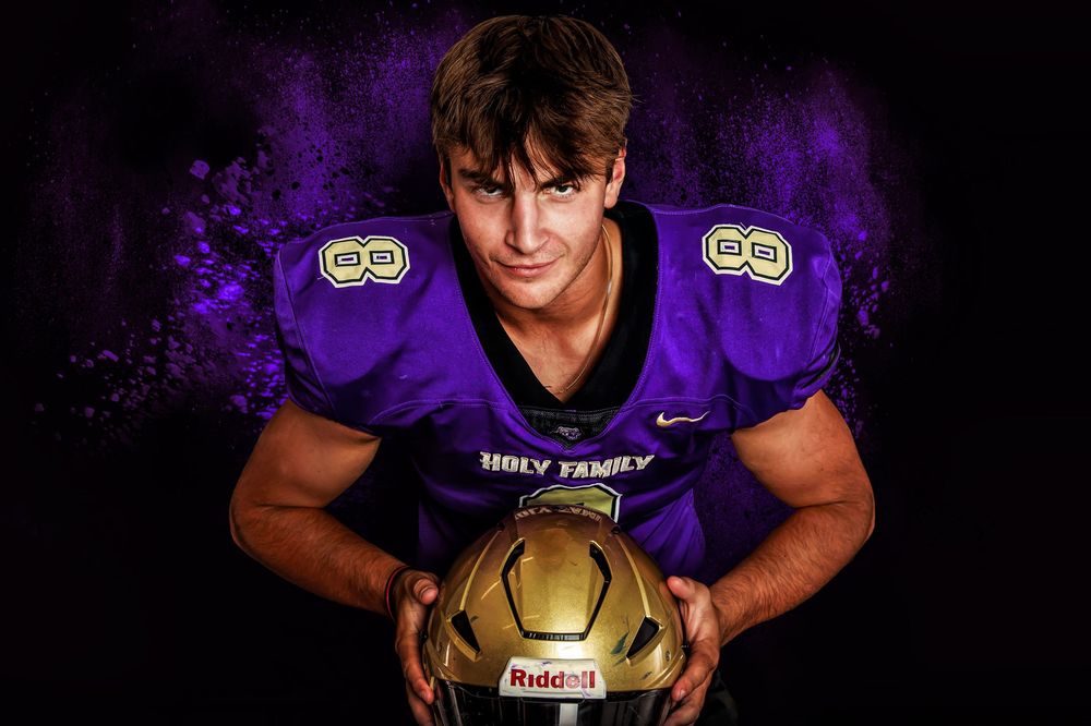 Player poses with football in purple and gold Holy Family uniform against dark backdrop.
