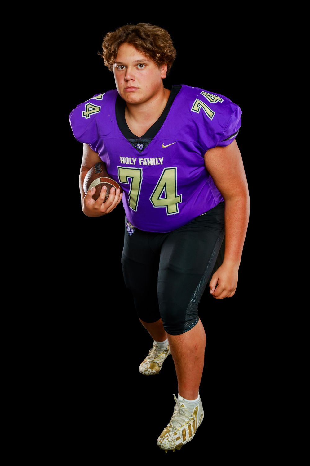 Football player in purple Holy Family jersey number 74 poses with ball against black background.