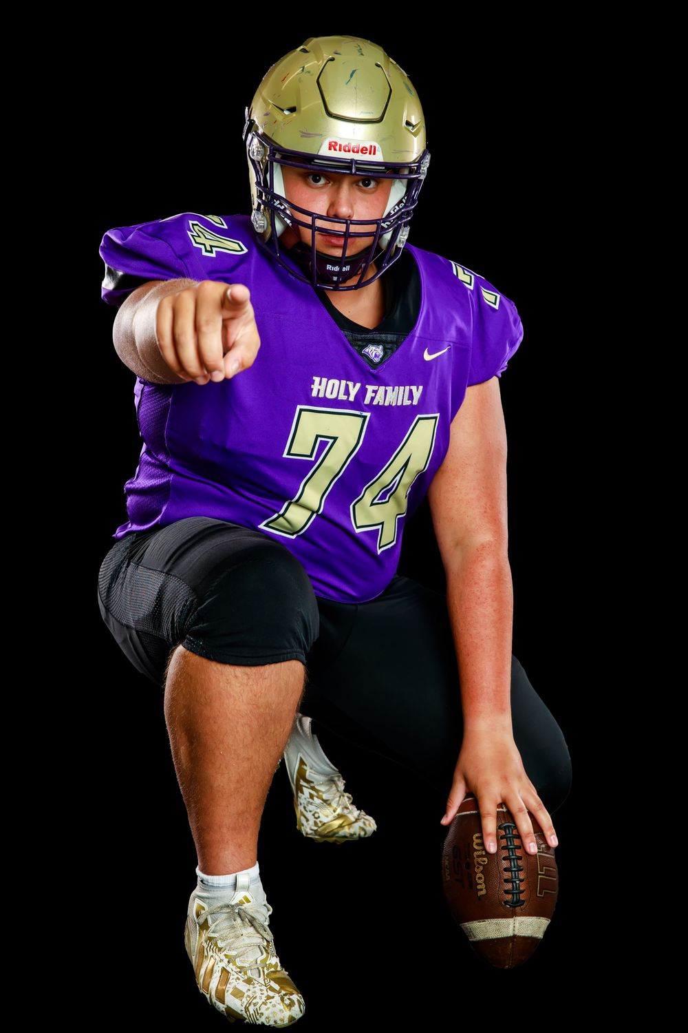 Football player in purple Holy Family uniform #74 poses with ball in athletic stance against black background.