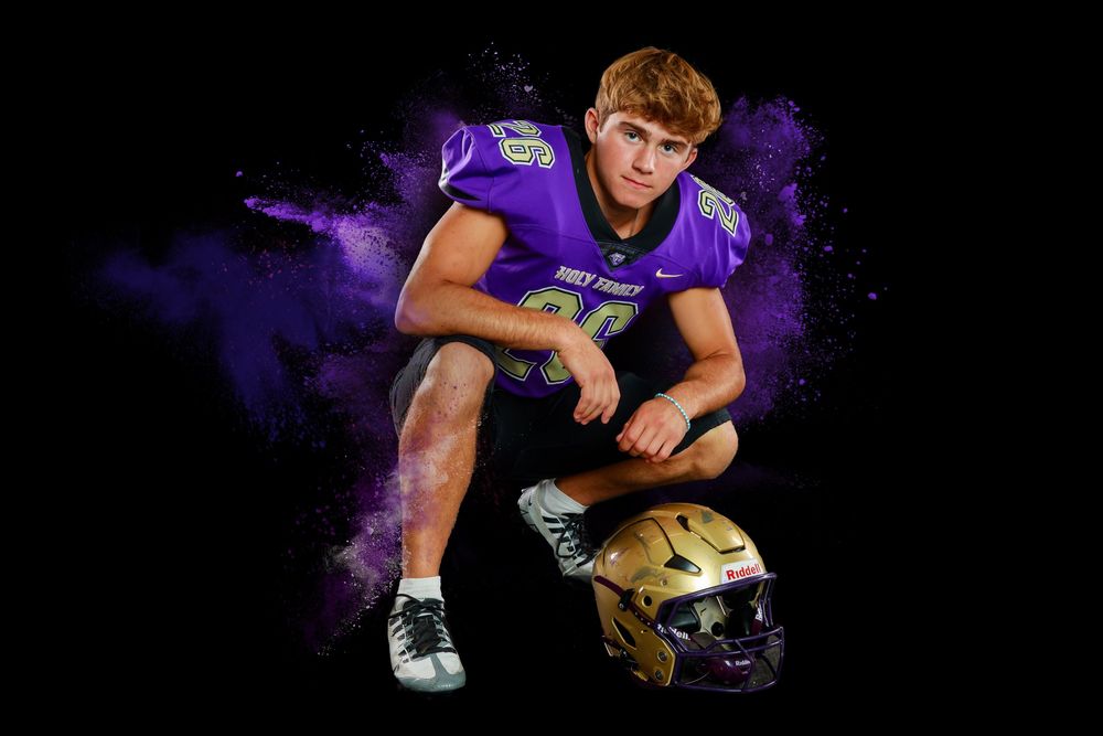 High school athlete in purple uniform poses with football helmet in dramatic lighting with purple smoke effect.