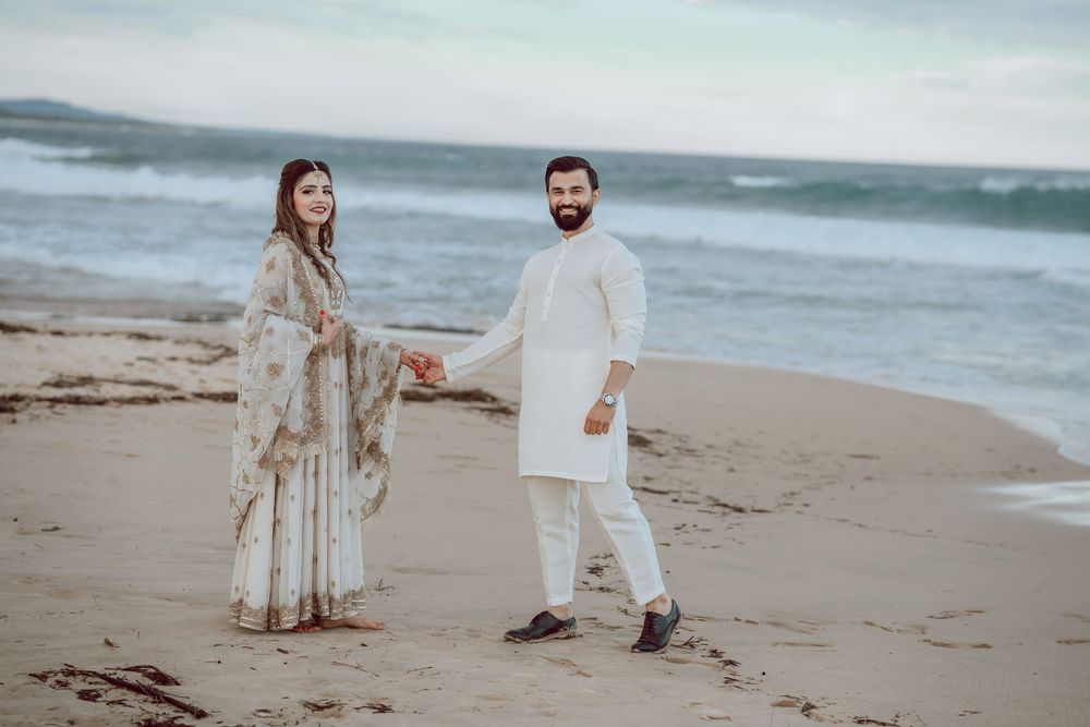 Two people walk hand in hand on a sandy beach during sunset wearing coordinated white and beige outfits.
