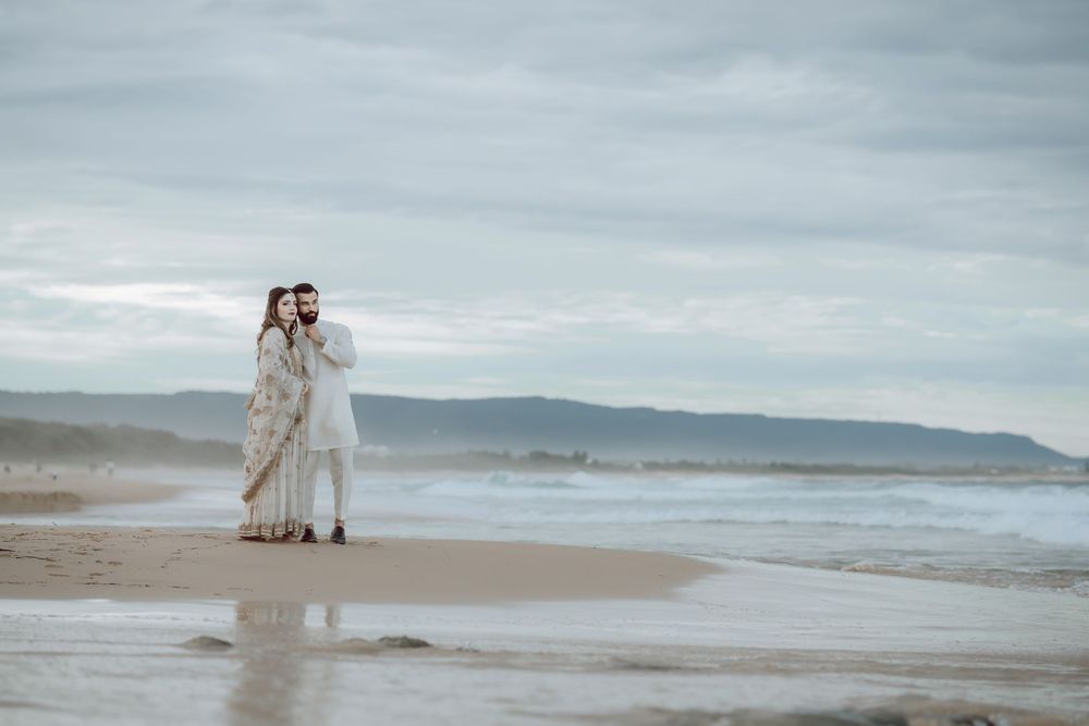 A romantic couple in white walking along a misty beach shoreline with rolling waves and cloudy skies in the background.