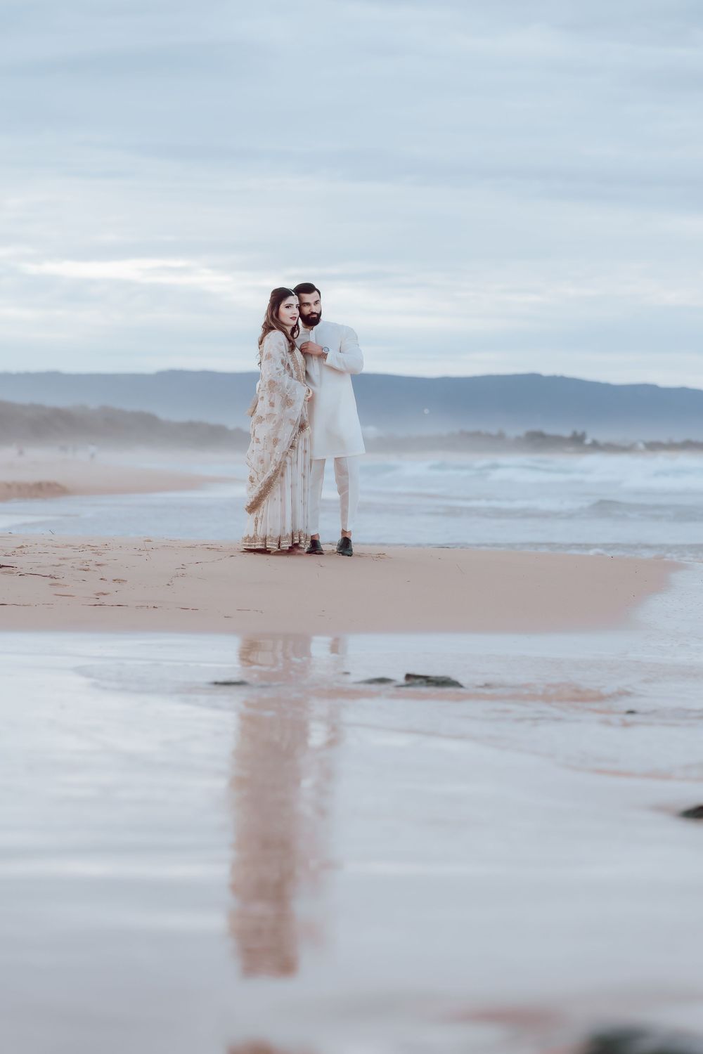 A couple walks hand in hand along a misty beach at sunset with mountains in the background.