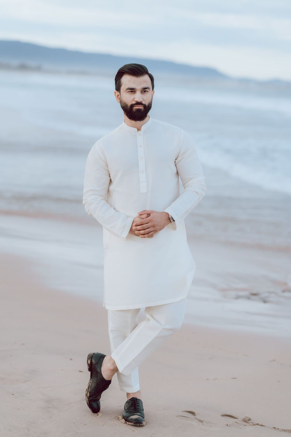 Person wearing white linen clothing poses thoughtfully on a misty beach at sunset.
