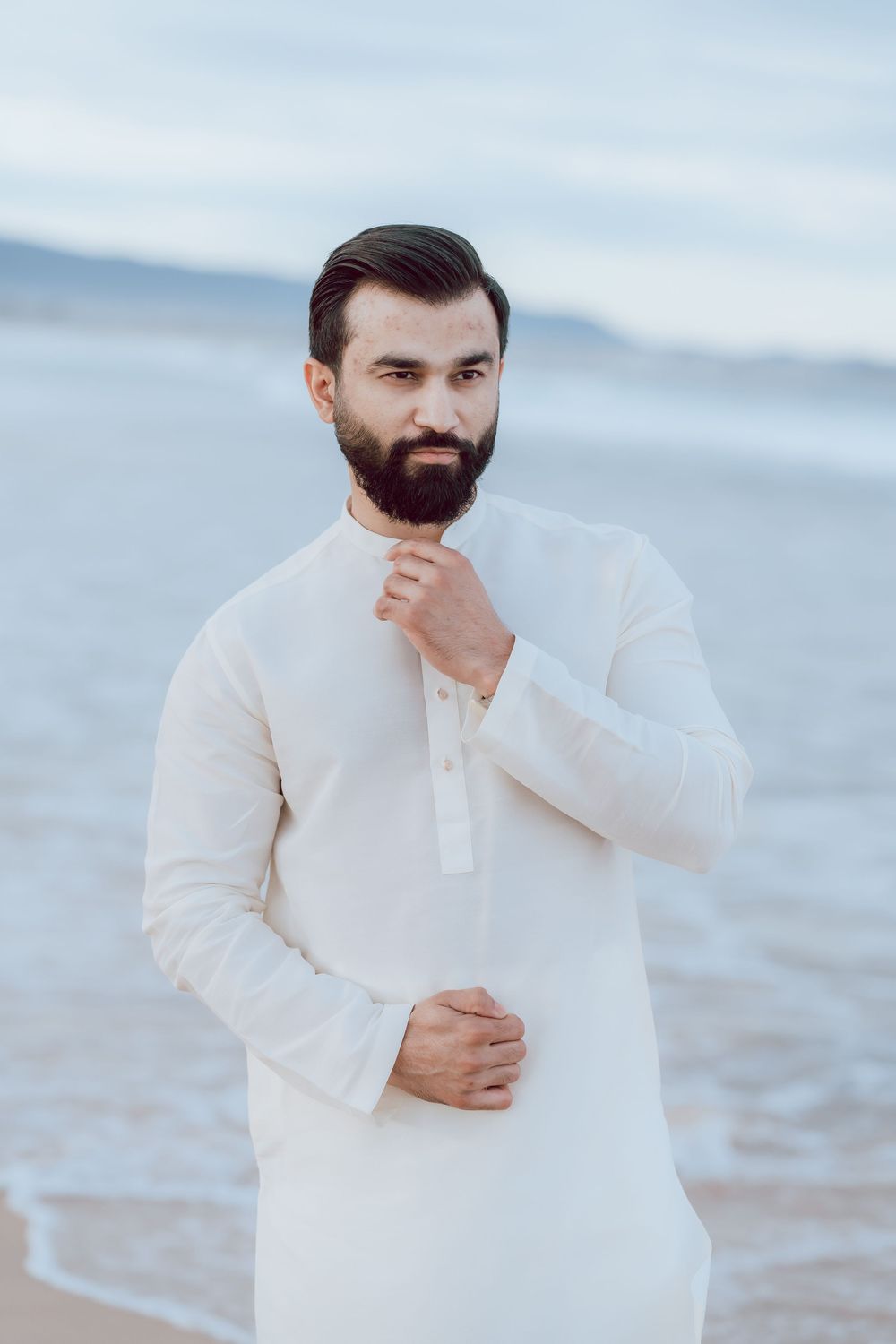 Person in white traditional kurta with dark beard stands thoughtfully by the ocean at dusk.