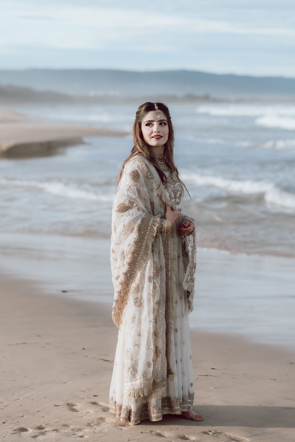 A figure in an elegant lace dress stands gracefully on a misty beach with rolling waves in the background.