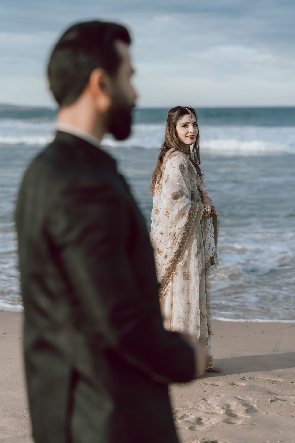 A couple stands apart on a moody beach with waves in the background as the woman wears a light floral dress.