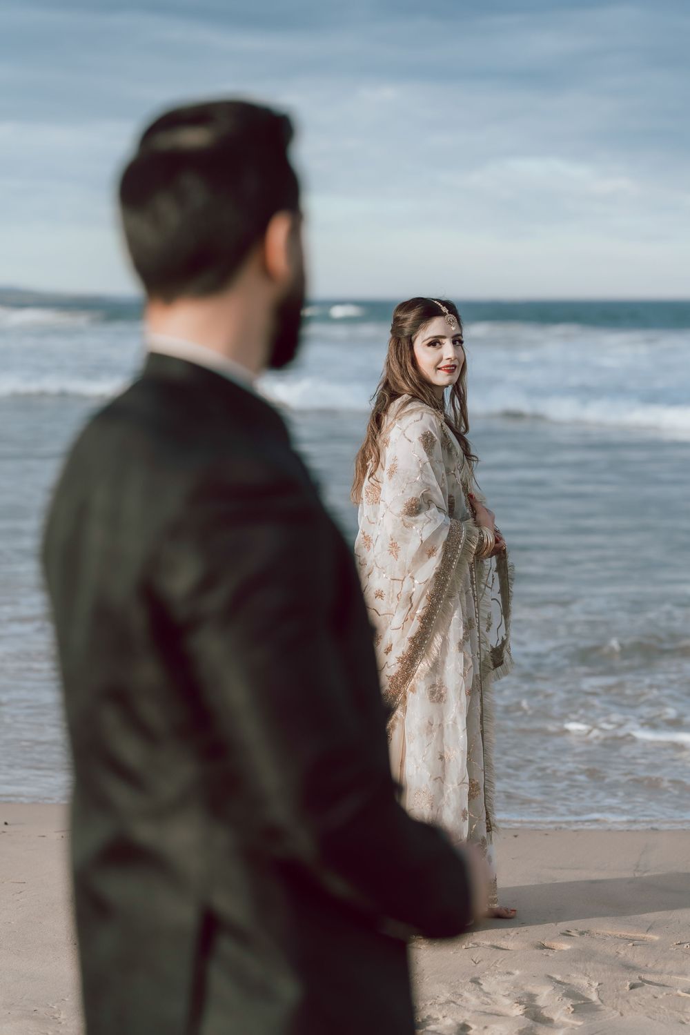A couple stands on a beach at sunset with the ocean waves and clouds creating a moody coastal atmosphere.