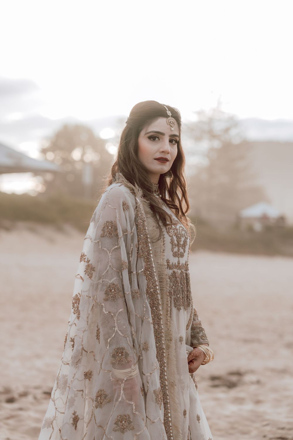 Ethereal portrait of person wearing ornate lace dress with floral embellishments standing in dreamy golden light.
