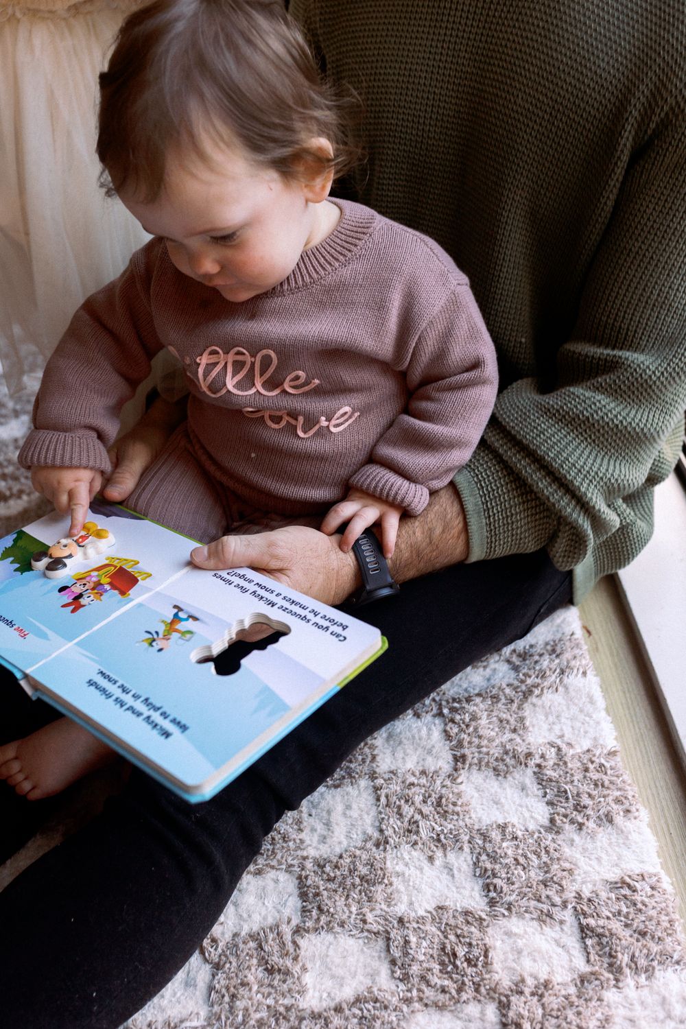 Child in a pink sweater reads a picture book while sitting on a patterned rug.