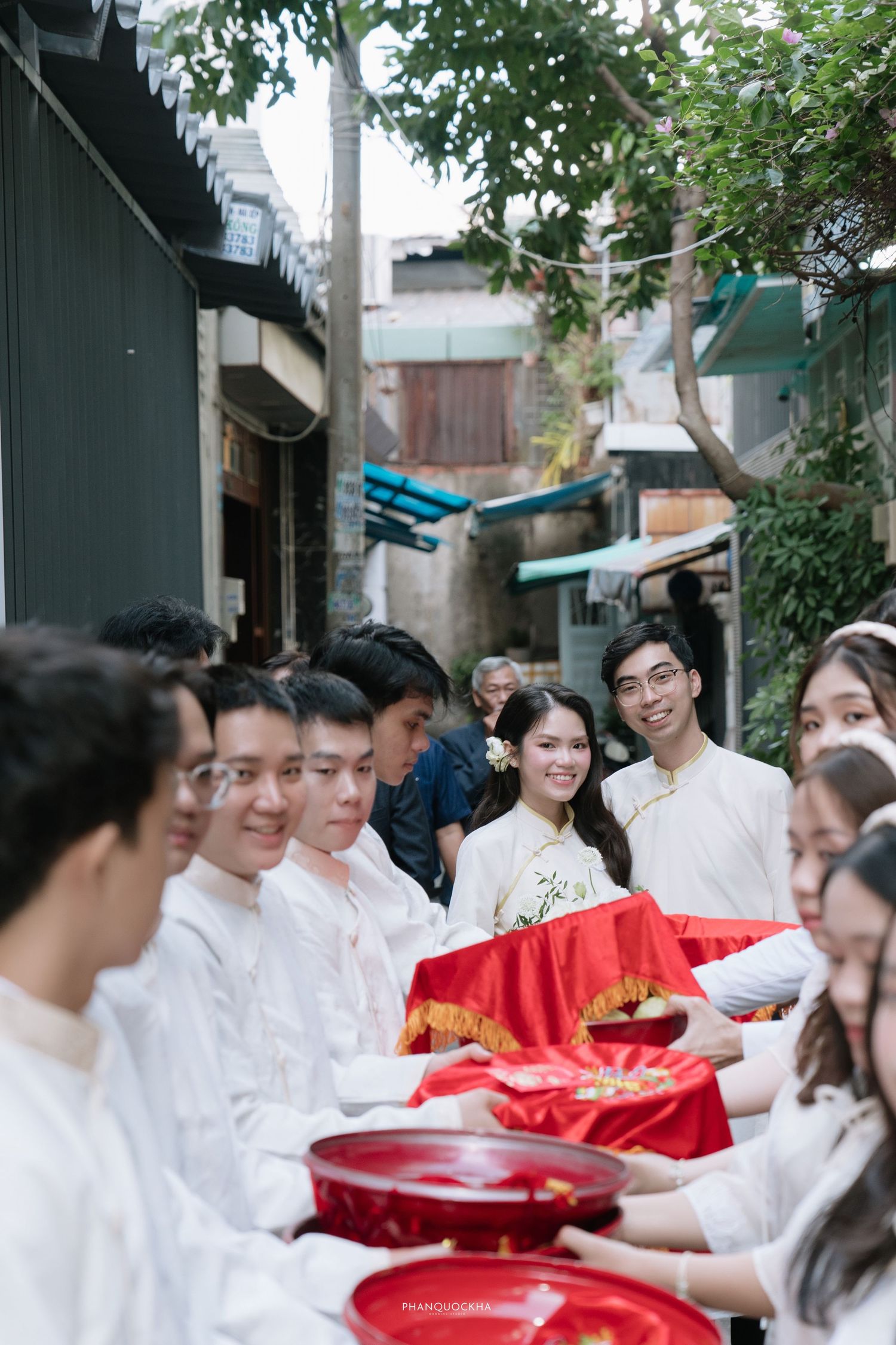 Vietnamese wedding procession with guests in white ao dai carrying red lacquer trays of gifts.