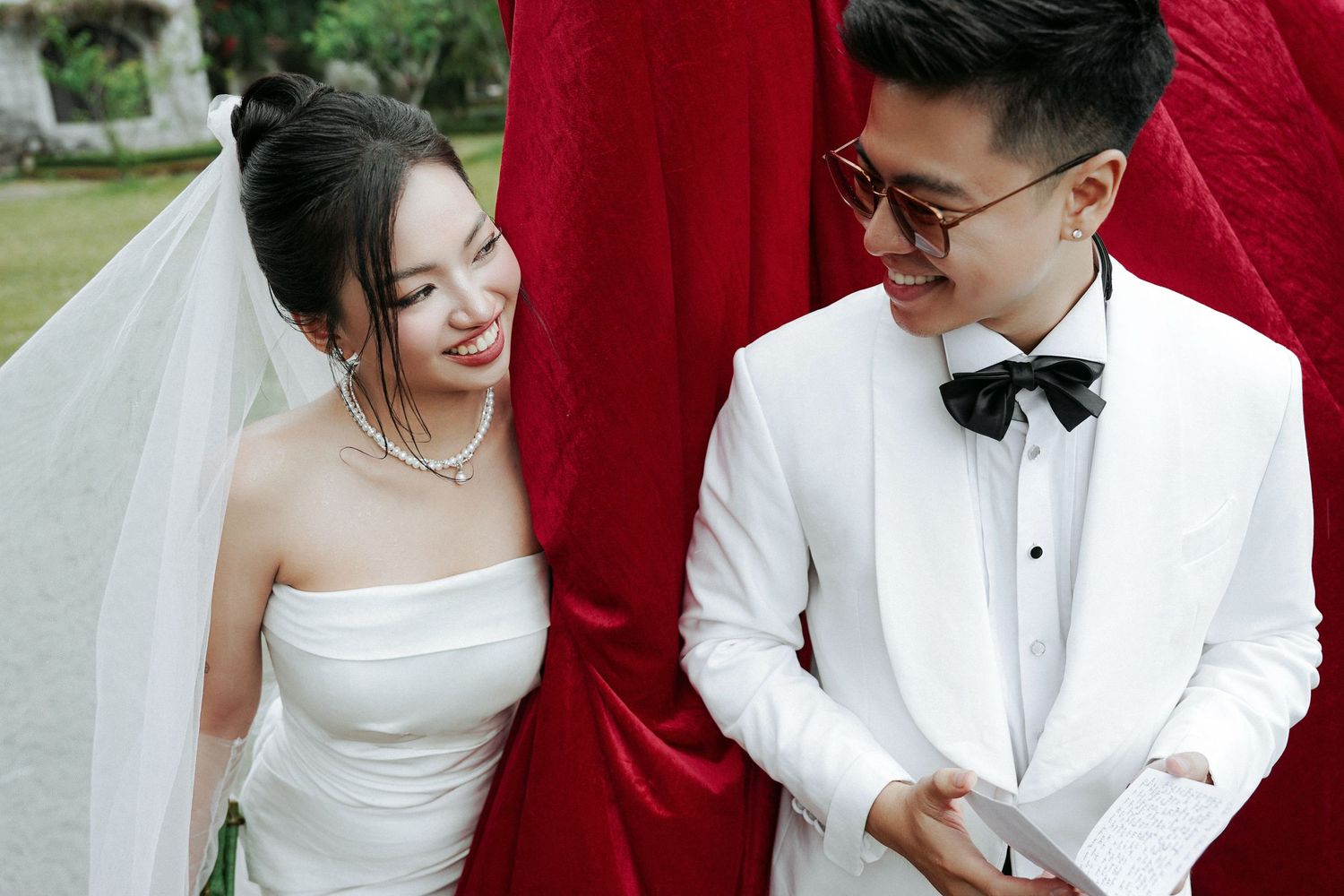 Smiling Asian bride and groom in white wedding attire share a joyful moment before red drapes outdoors.