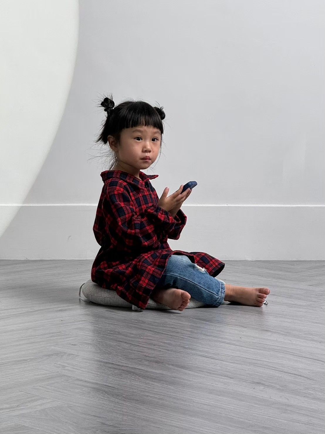 Young child in red plaid shirt and jeans sits on gray floor looking at mobile phone in minimalist setting.