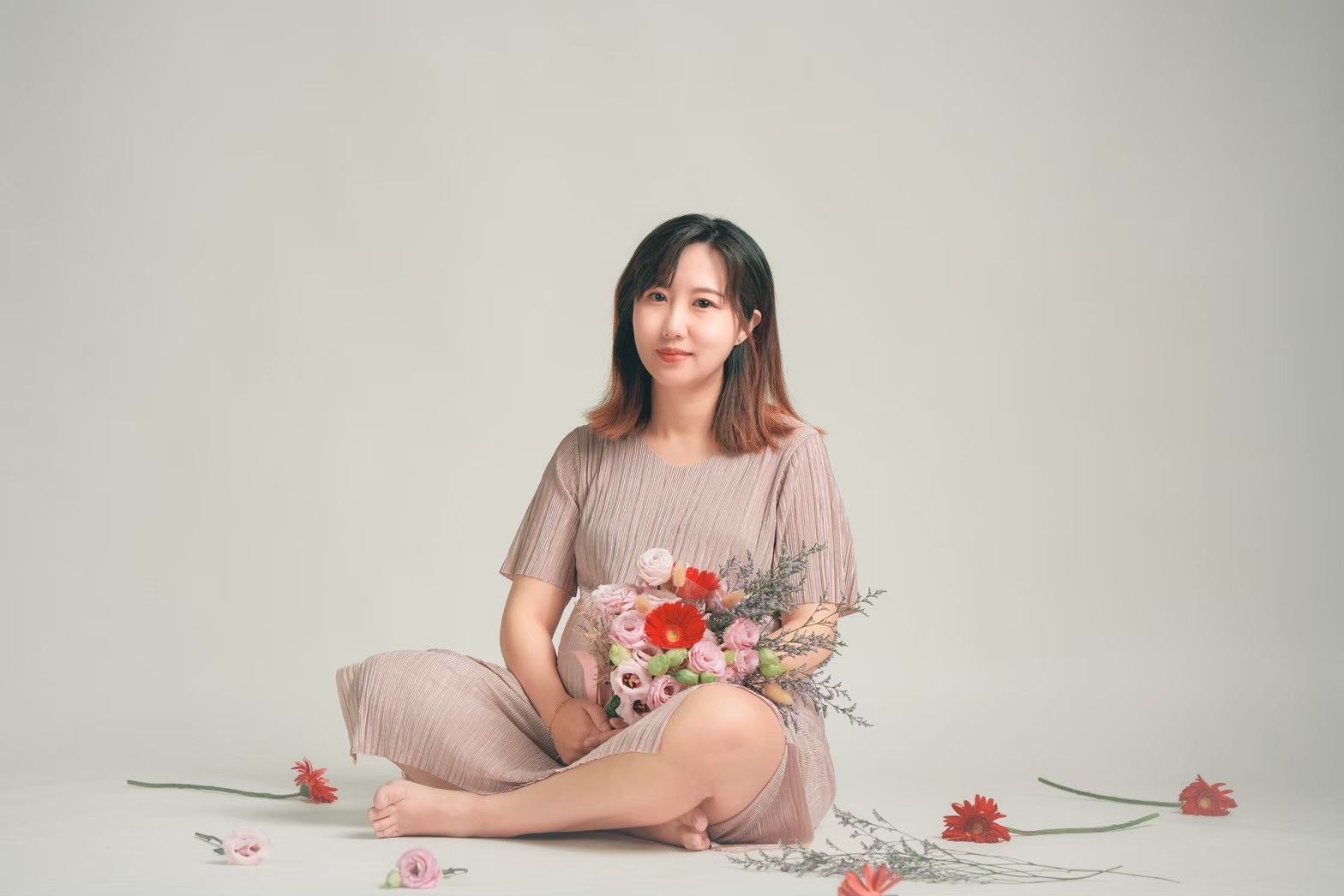 Woman in mauve dress sits elegantly with fresh flowers and teacup on light neutral background.