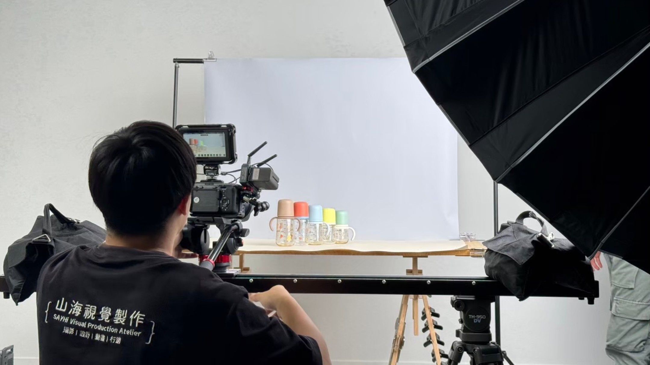 Camera operator films product bottles on set during commercial video production shoot against white backdrop.