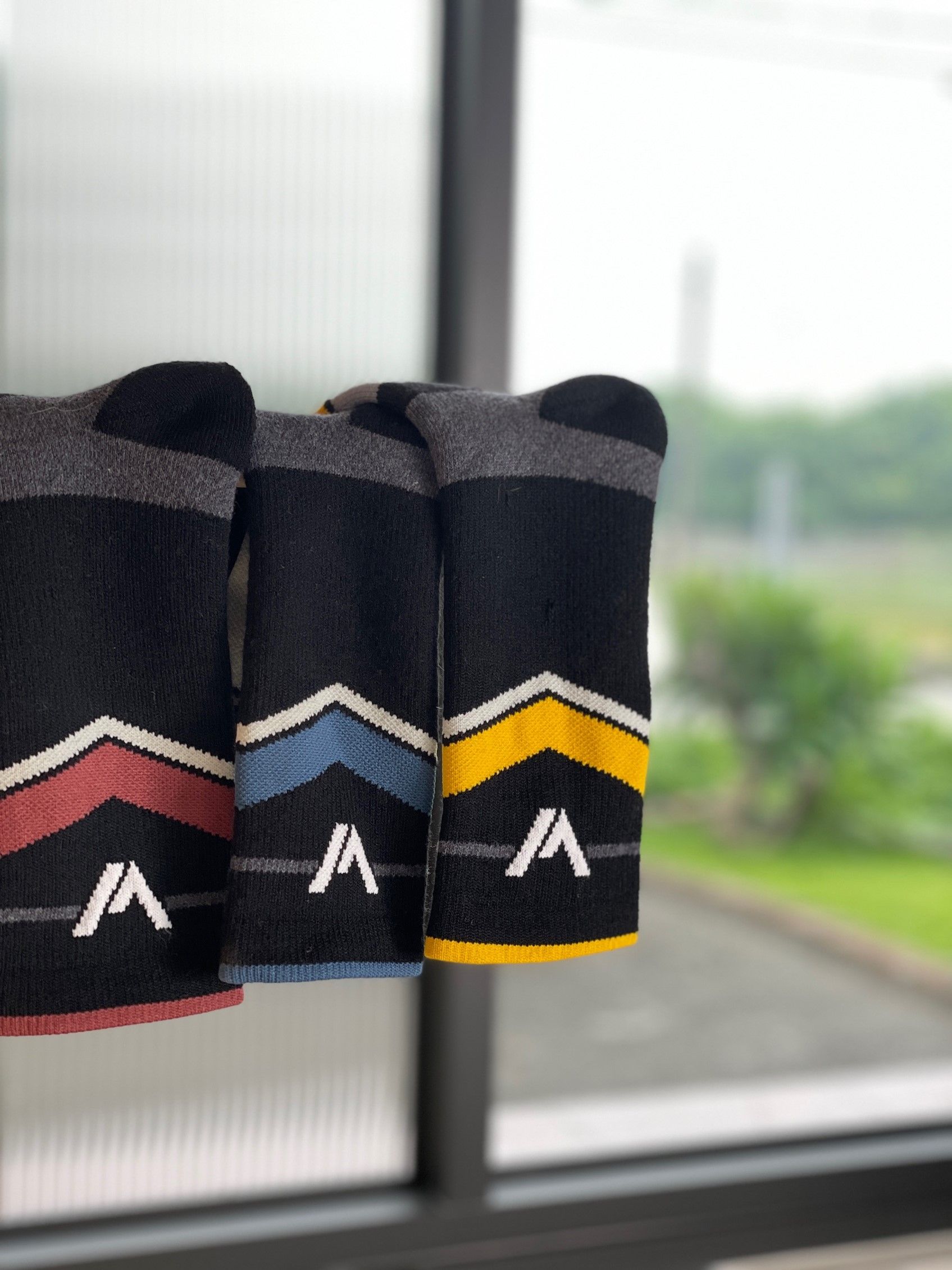Three athletic socks with stylish stripes in black, gray, and colored accents featuring the letter A logo displayed by a window.