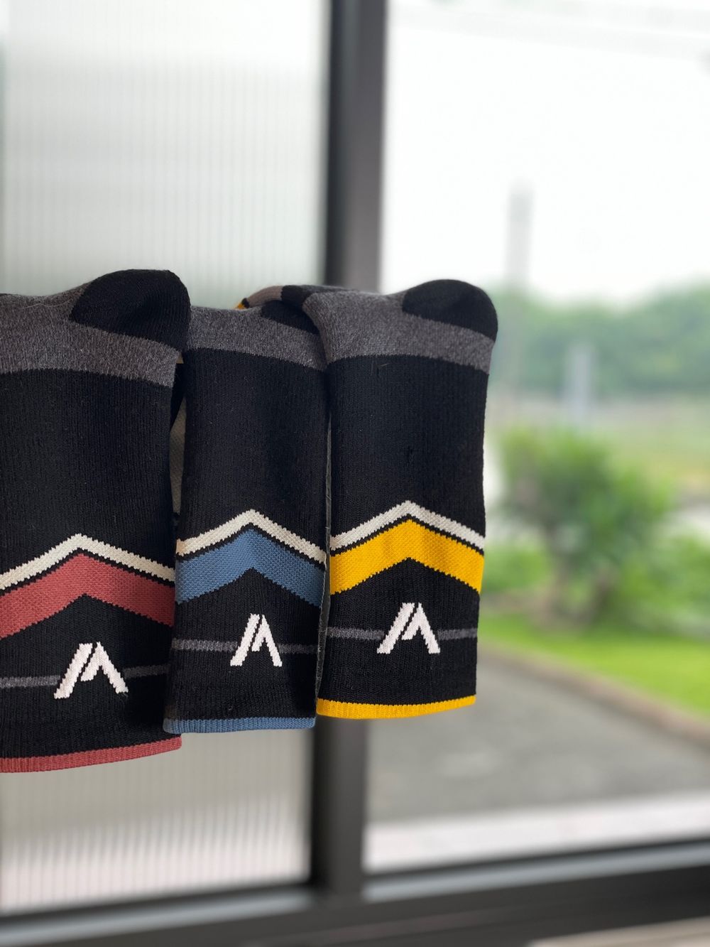 Three athletic socks with stylish stripes in black, gray, and colored accents featuring the letter A logo displayed by a window.
