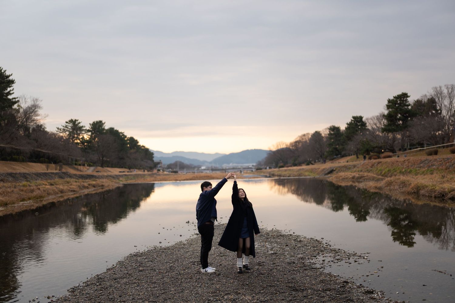 Couple holding hands up on a scenic riverbank at sunset with mountains reflected in calm water.