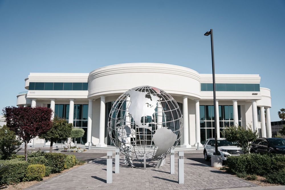 Modern white architectural building exterior featuring curved walls, glass windows and landscaping under blue sky.