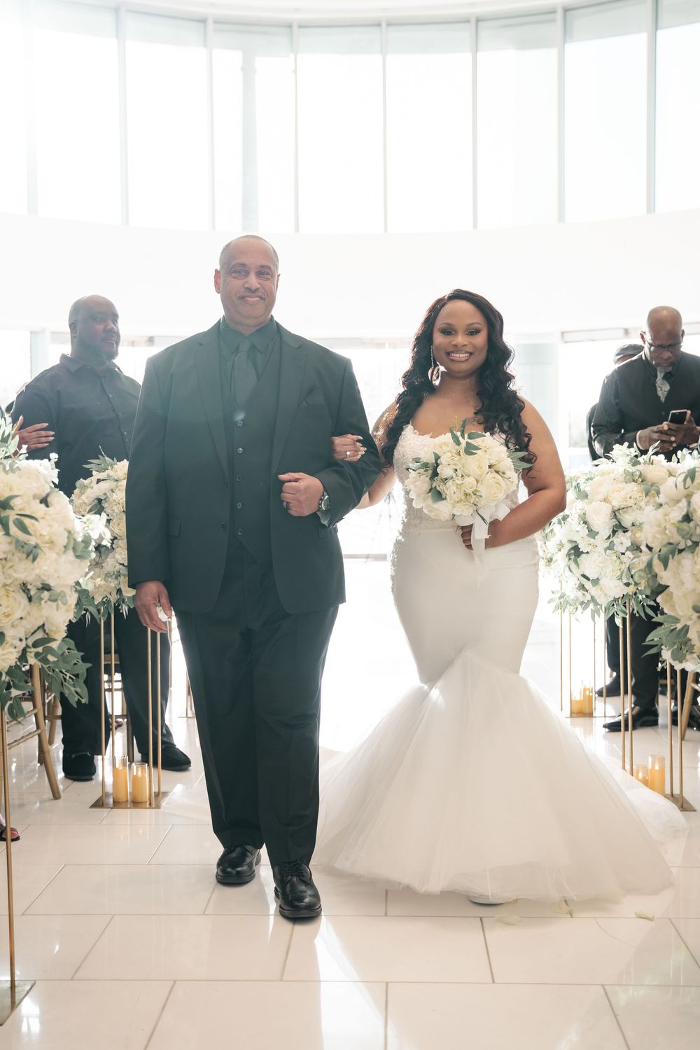 Wedding party members in black attire stand alongside white decor in an elegant bright venue.