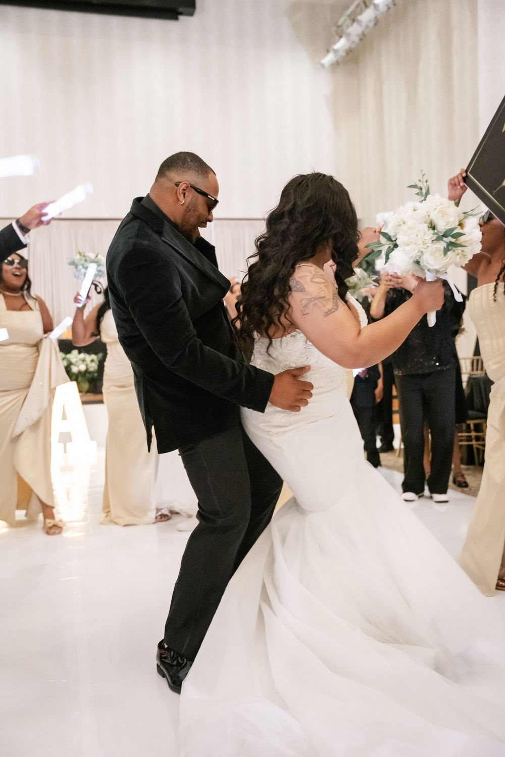 A couple dances together at their wedding reception on a white dance floor surrounded by guests and floral decorations.