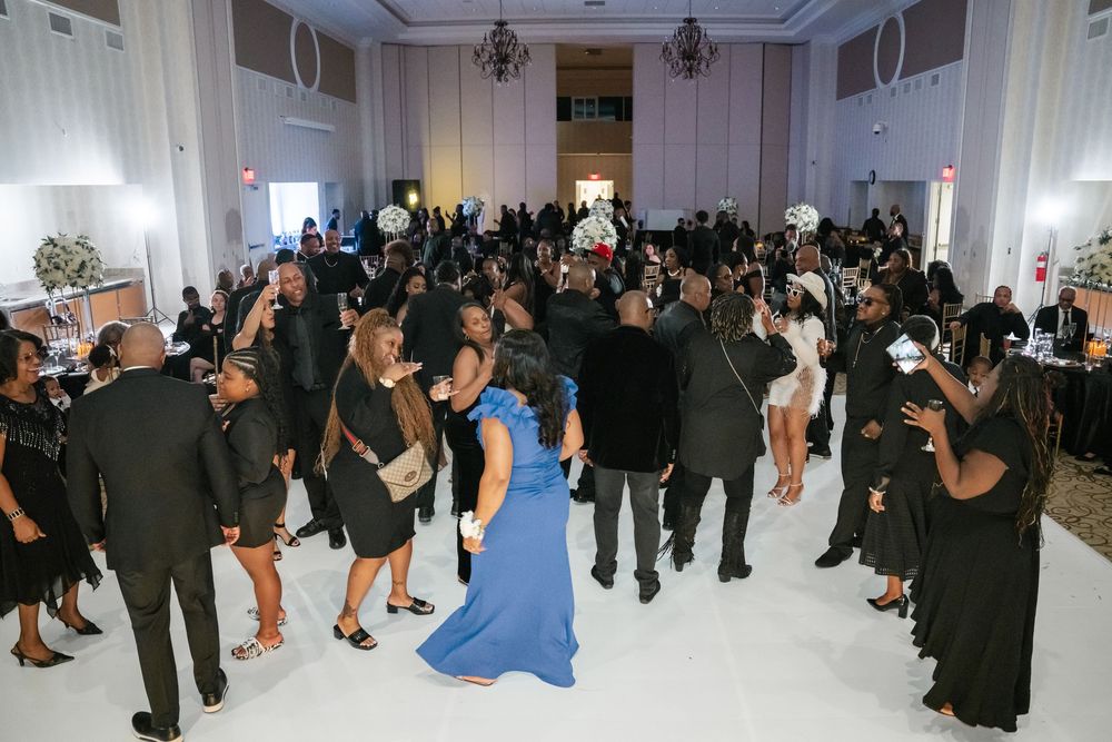 Party guests dressed in formal black attire dance together in an elegant white reception hall.