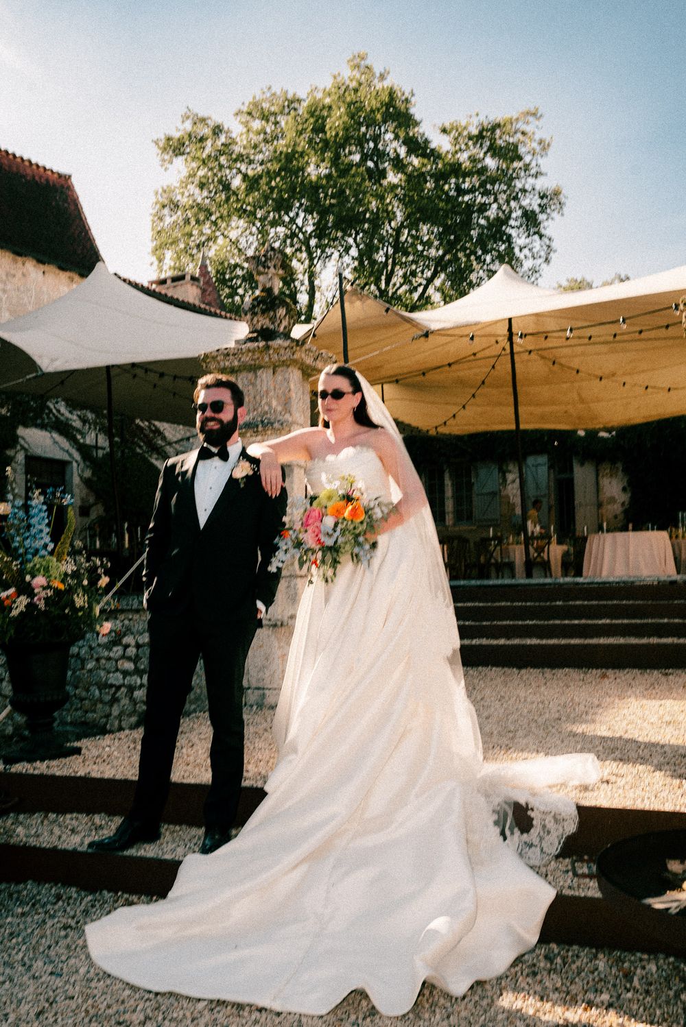 A wedding couple walks hand in hand together outside on a sunny day with their flowing white dress trailing behind.