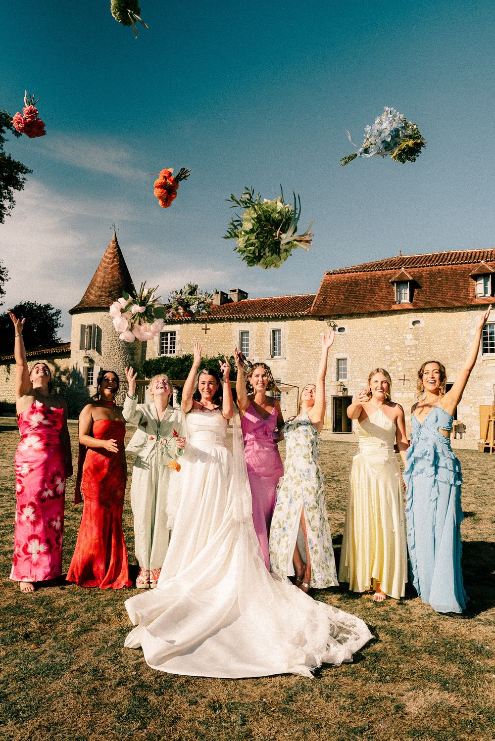 Wedding party lined up outdoors with colorful bouquets being tossed in the air against a French chateau backdrop.