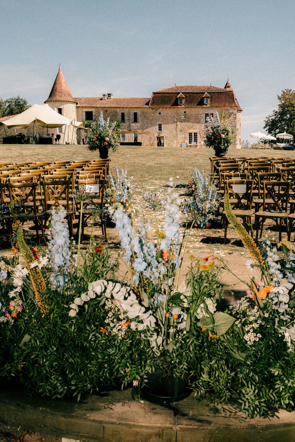 White delphinium flowers bloom in a rustic garden with stone buildings and wooden chairs arranged in the background.
