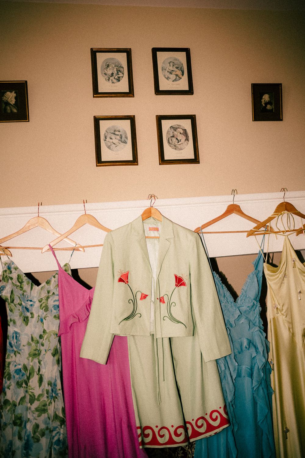 Clothing hangers display an assortment of pastel dresses in mint green, turquoise, and pink against a beige wall with framed art.