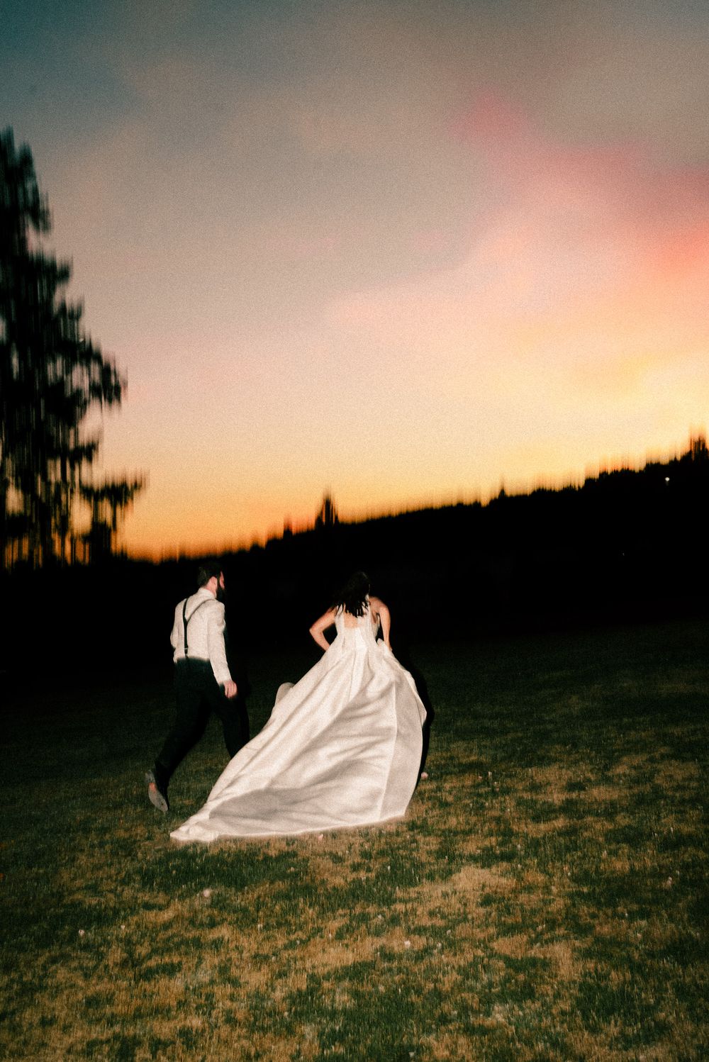 A flowy white wedding dress billows dramatically across a hillside field at sunset creating an ethereal silhouette.