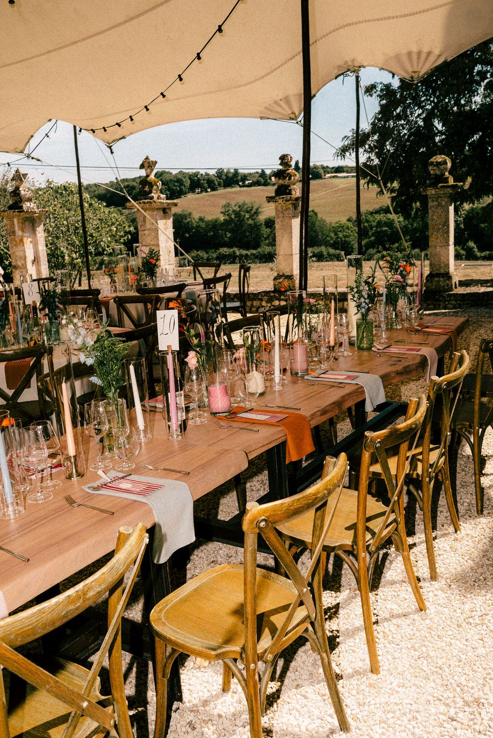 Long dining tables set outdoors with wooden chairs and hanging glass decorations under tent canopies for an elegant event.