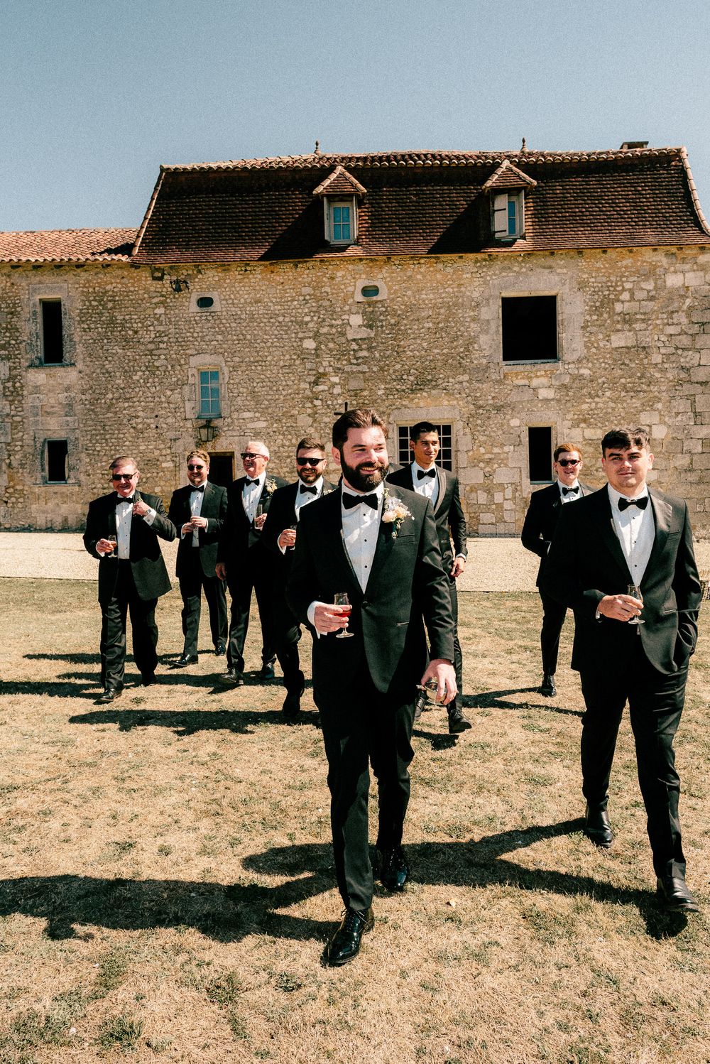 Wedding party in black tuxedos walking across open countryside by rustic stone buildings in daytime.