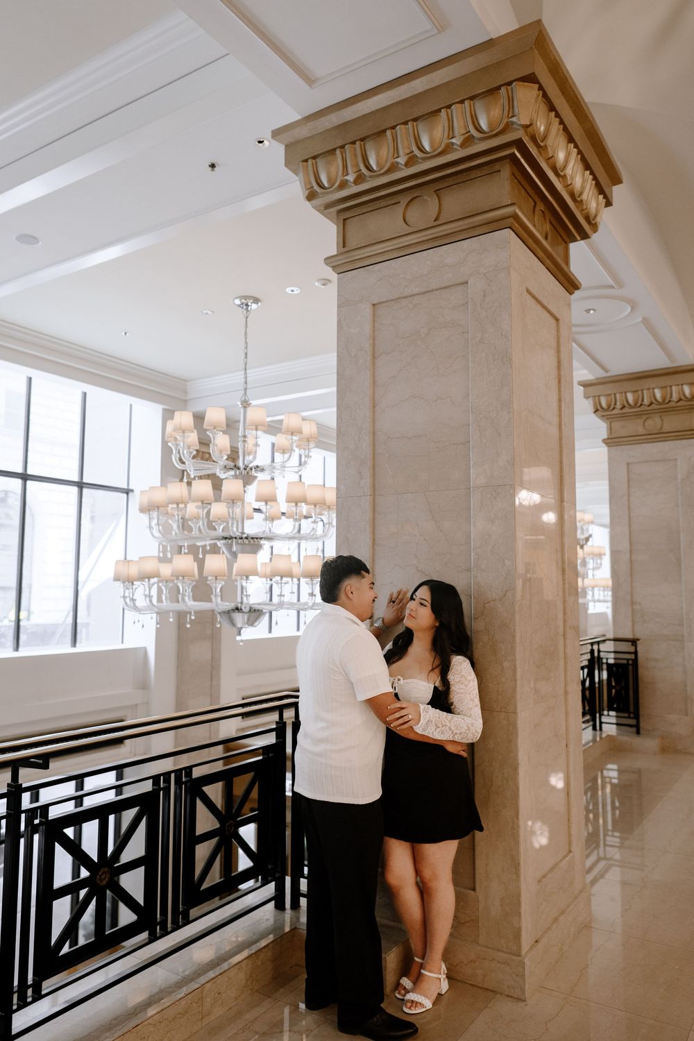 A bright interior hallway with marble columns, black railings and chandeliers showing a couple in contrasting outfits.