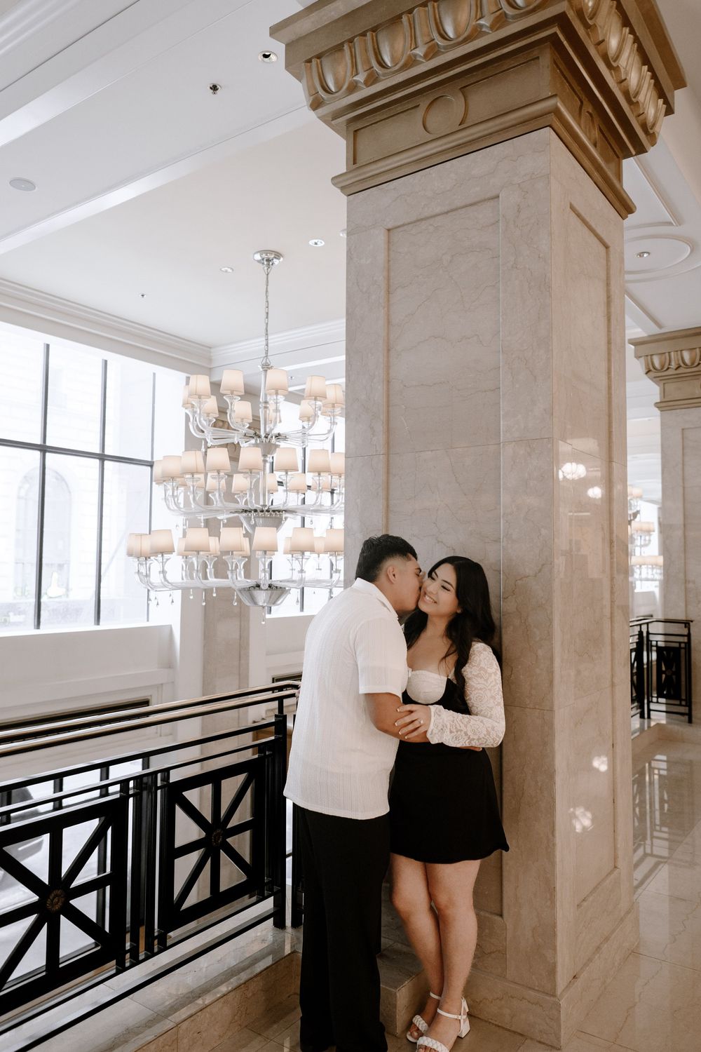 A couple shares romantic moments in an elegant hallway with marble columns and crystal chandeliers.
